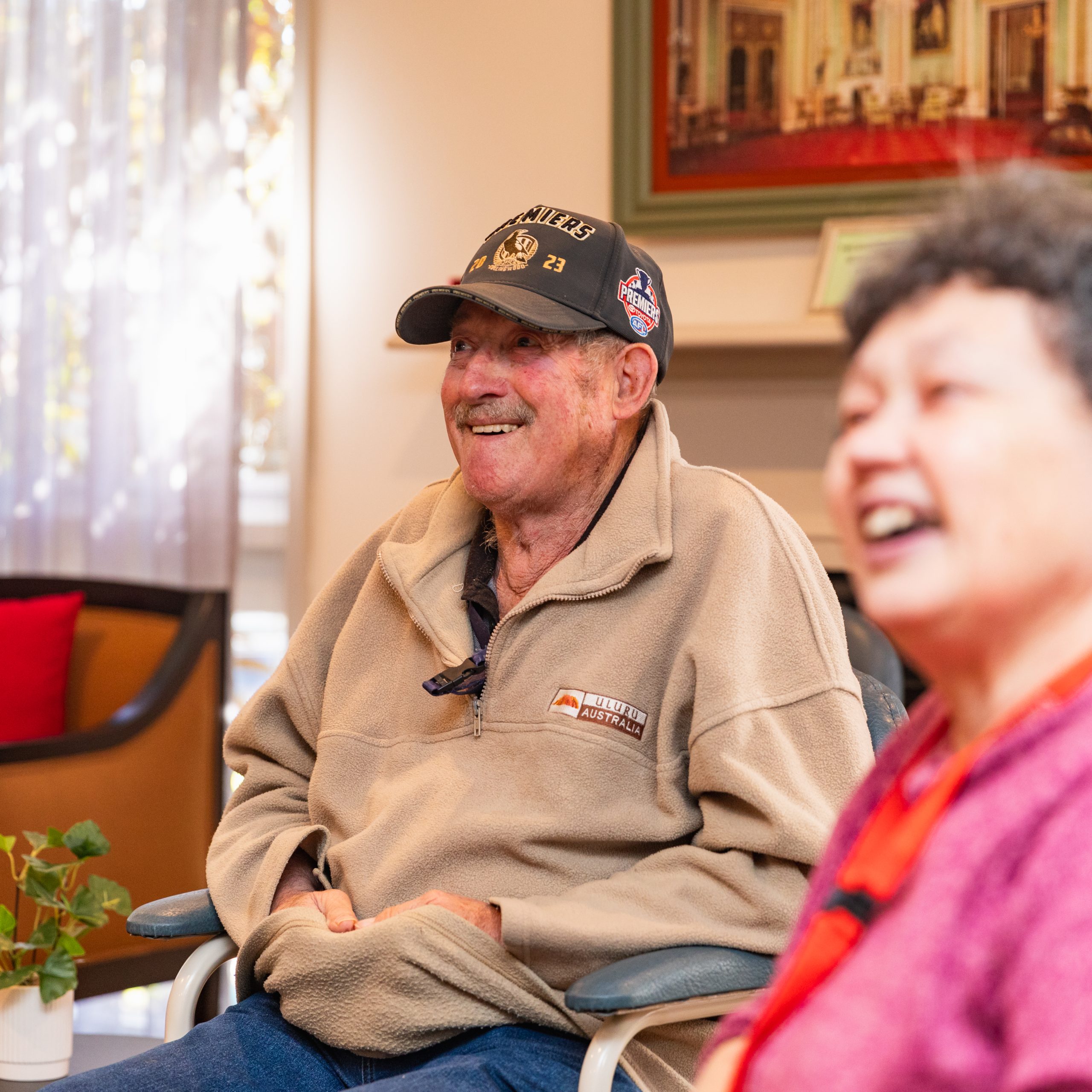 An older man with a baseball cap on smiling and an out of focus woman smiling in the same direction