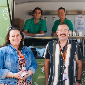 Four people at a food truck: two staff in green shirts inside, and two customers outside.