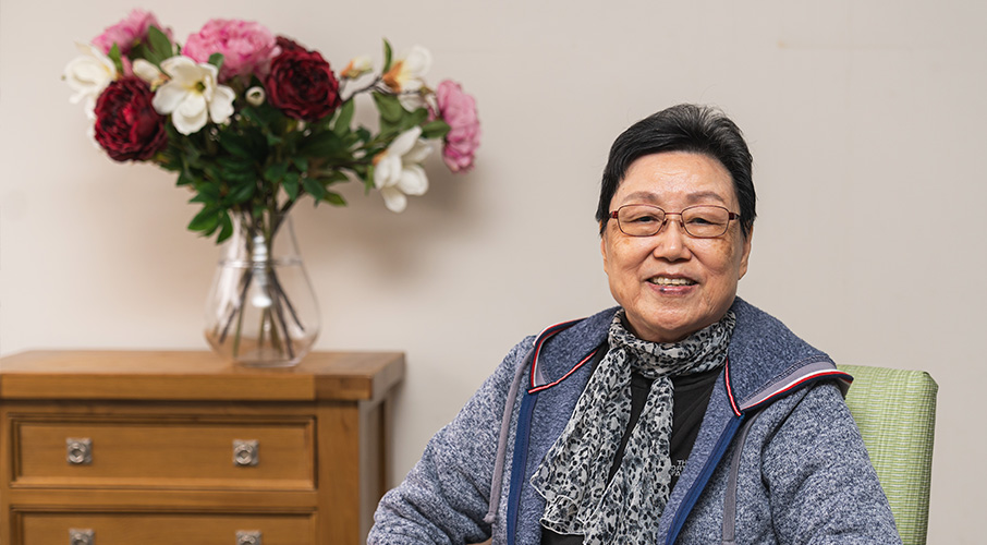 A woman sitting near a vanity chest that has bright pink flowers in a vase sitting on it.