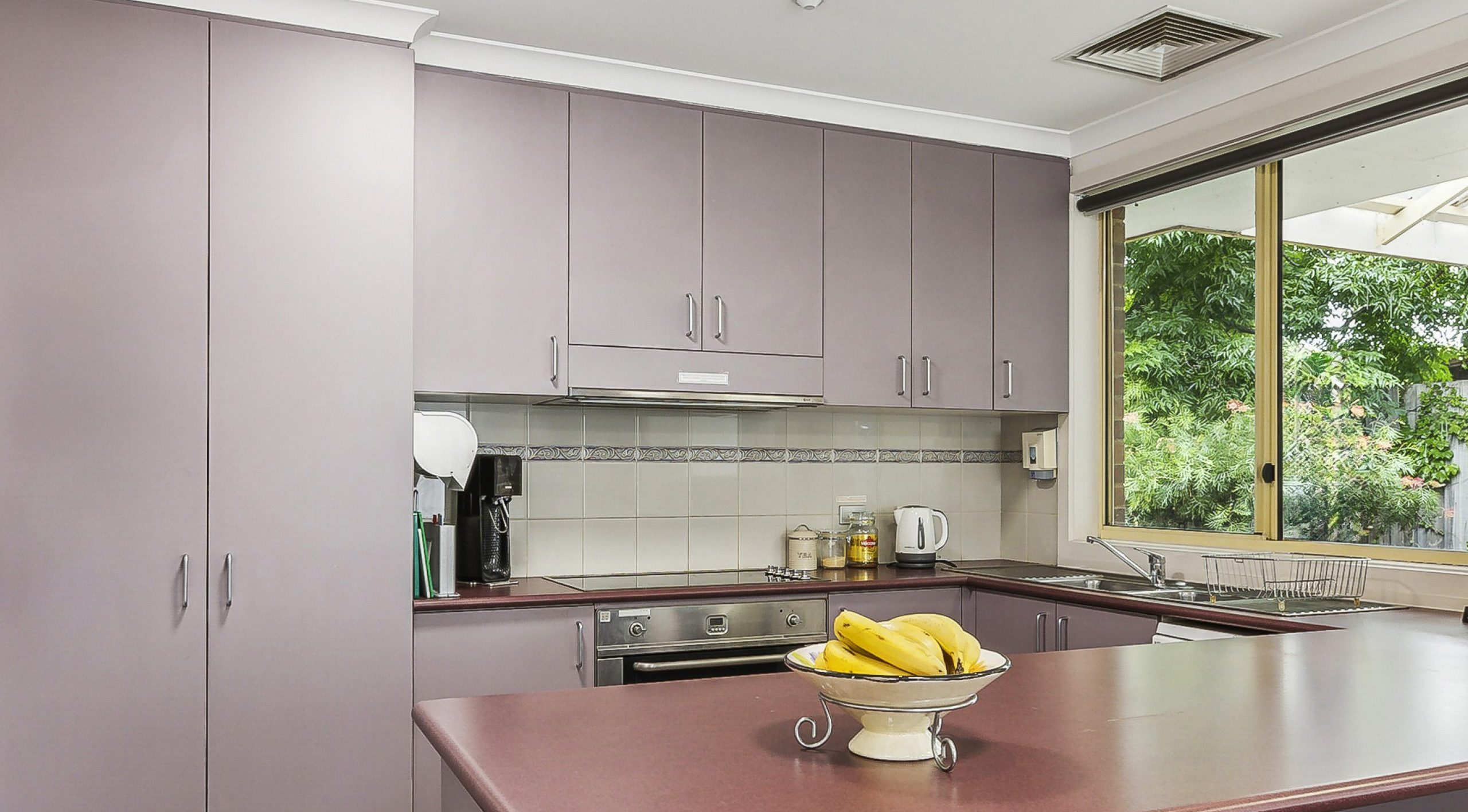 A modern kitchen interior with a focus on clean lines and functionality. The kitchen features a combination of white and lavender cabinetry, with stainless steel appliances including a stove, microwave, and refrigerator. A central island with a pink countertop holds a bowl of bananas, emphasizing the room’s lived-in feel. To the left, there is a fire extinguisher mounted on the wall next to the refrigerator. A wall clock above the microwave indicates the time. The space is well-lit by ceiling downlights and natural light from an unseen window