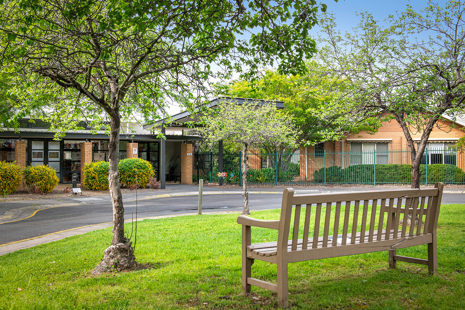 A bench facing a building entrance with green trees around.