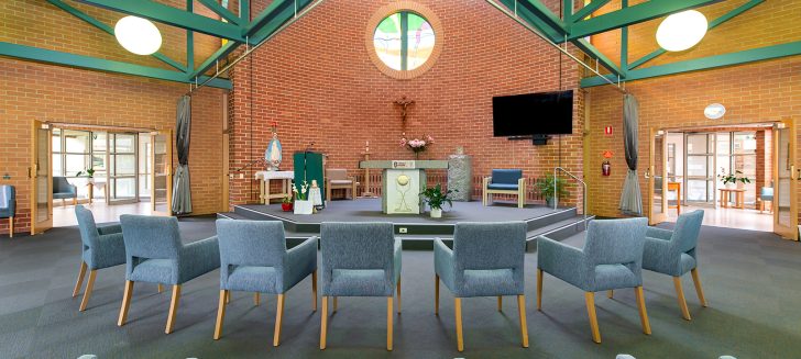 A chapel with green beams across the ceiling. Small armchairs are arranged to face the altar.