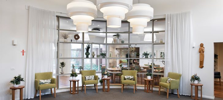 A living space with green armchairs along the window which looks into the dining room. A large light feature hangs in the centre of the room.