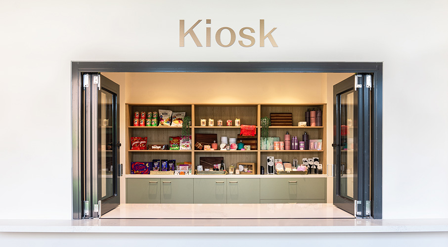 A shop opening with the word 'kiosk' above it on silver. Behind a shop bench is shelves filled with products.