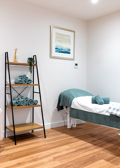 A tidy spa room with a massage table, neatly folded towels, and a decorative shelf against a white wall. A framed artwork hangs above the table.