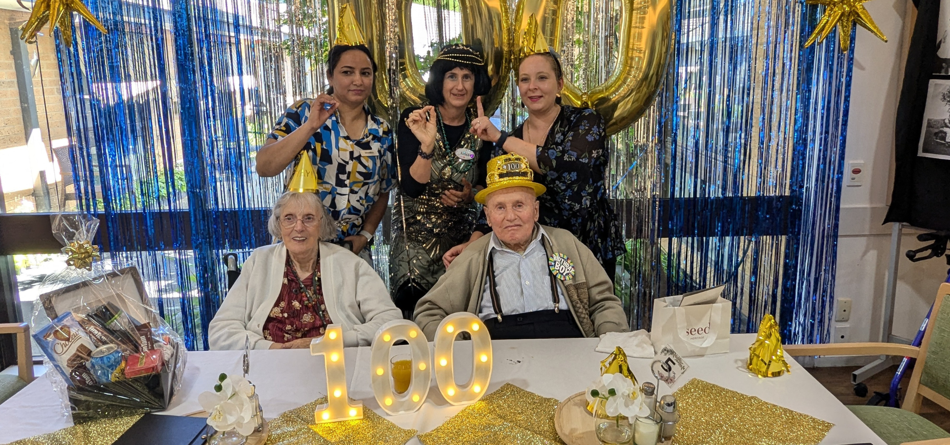 An older man and woman sit at a table with a 100 sign in front of them. Three woman stand behind signalling 100 with their hands.