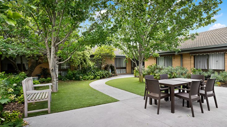 A paved path through a garden with grass and trees, an outdoor table and chairs sits near the grass.