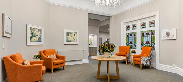 A living space with orange armchairs along the wall. A chandelier hangs from the middle of the ceiling with a coffee table below it.