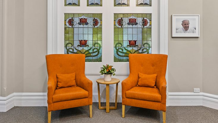 Two bright orange armchairs with a small side table between them and a stained glass mosaic behind them.