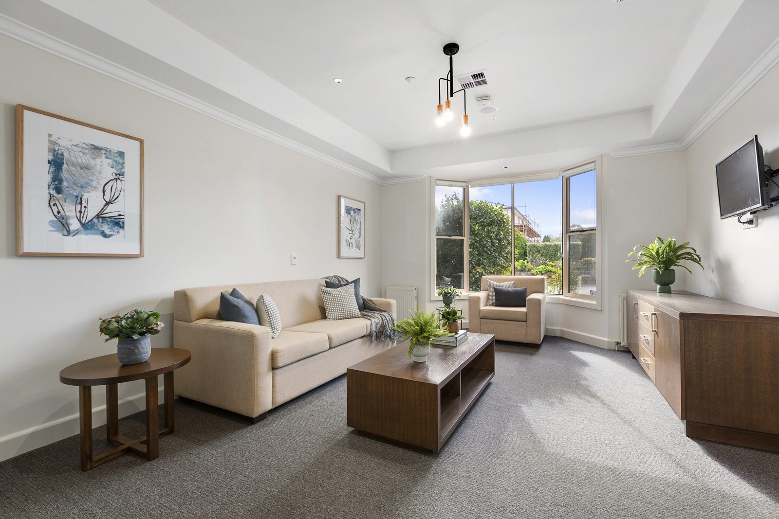 A sunlit narrow room with a cream three seater couch and matching armchair. Solid brown timber coffee table and matching side table next to the three seater couch with plant in a pot on it. Tv is on the wall above a dark wooden TV unit.