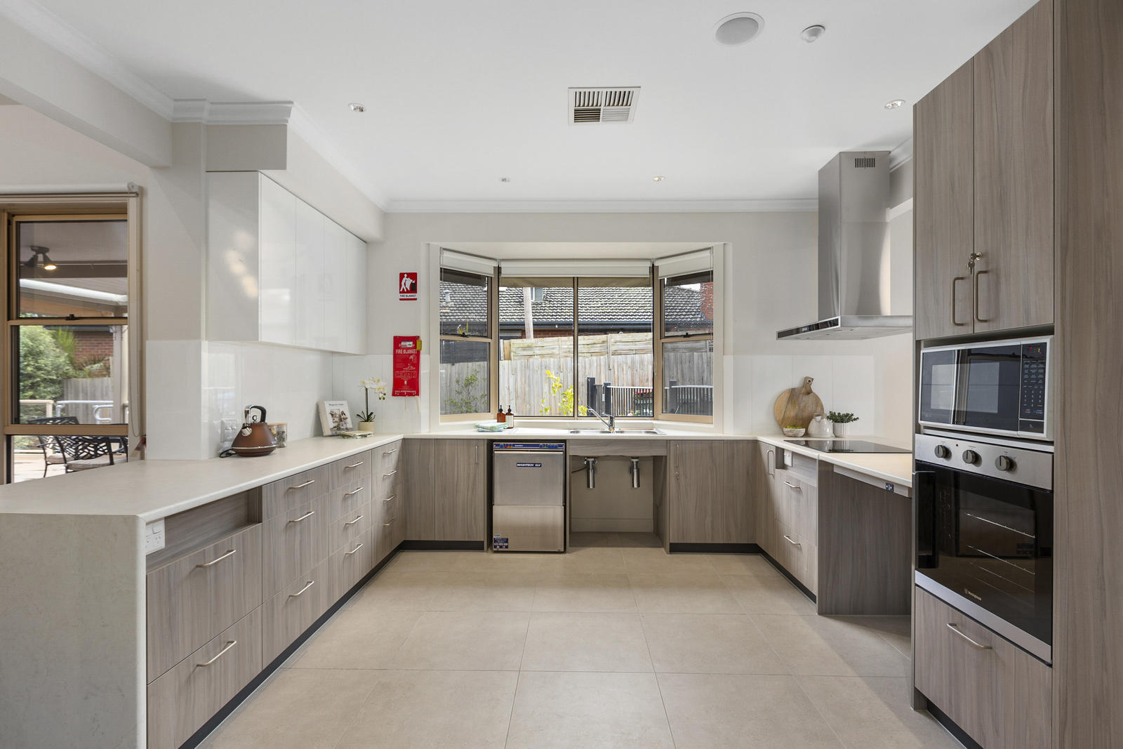 U shaped kitchen with light grey cabinetry. large window facing out to garden with a sink and silver dishwasher underneath it. Cabinet on right with microwave and oven set into it.