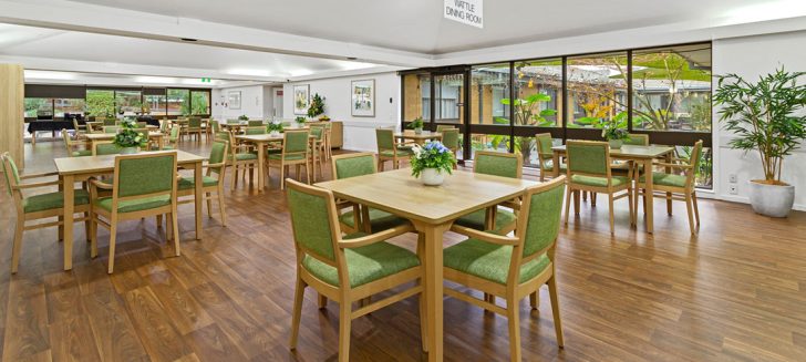 A spacious, lightfilled dining room with wooden square tables and green chairs.