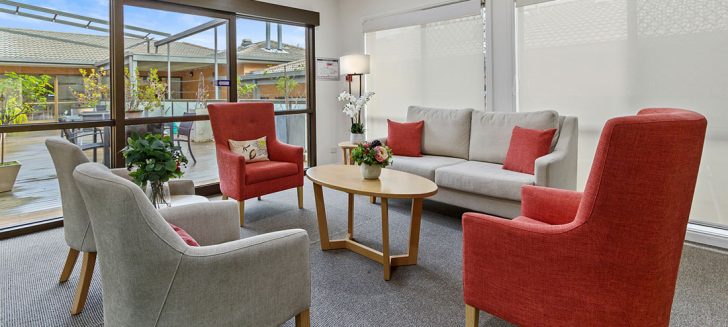 A grey couch against a window and 4 arms chairs. View out to a deck.