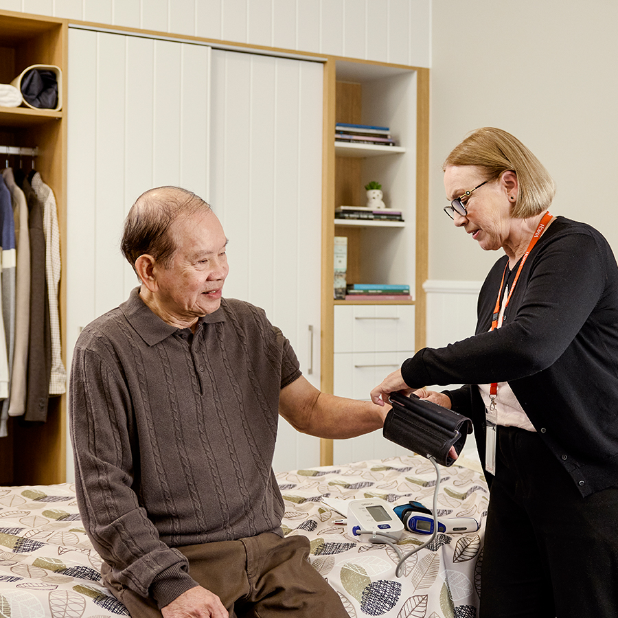 An older man sitting in his bedroom while a nurse takes his vitals as part of the EOL pathway.