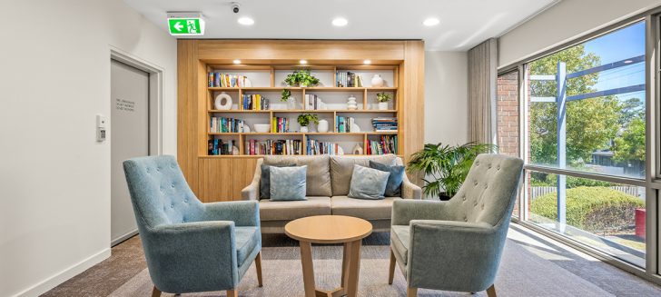 A living room with a filled book case and comfy chairs at Corpus Christi Clayton Aged Care.