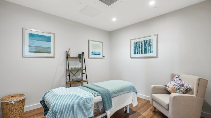 A massage room with a massage bed, paintings on the wall and a cream armchair.