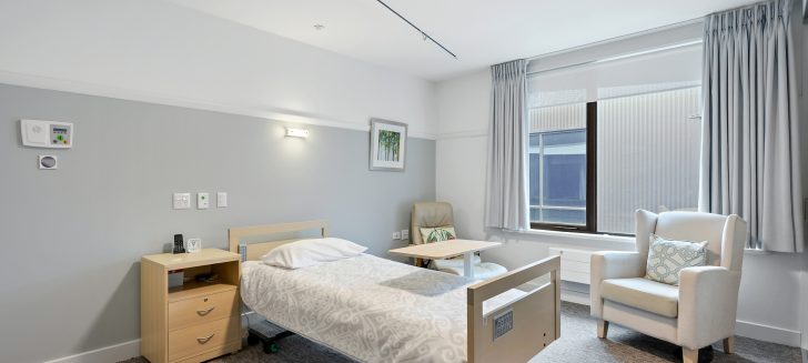 A bedroom with spacious windows, a bed with cushions and a chair in the corner.