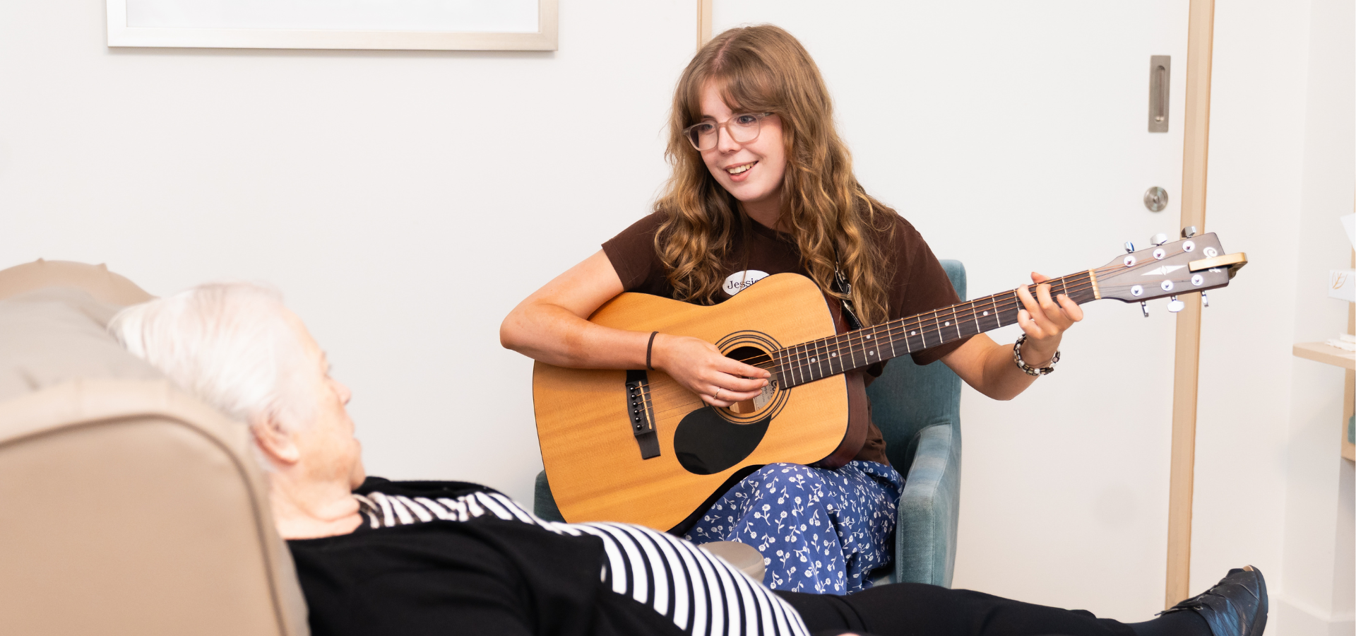A woman holds a guitar and sings to an older lady lying down