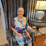 An older woman sits on a chair. She is smiling.