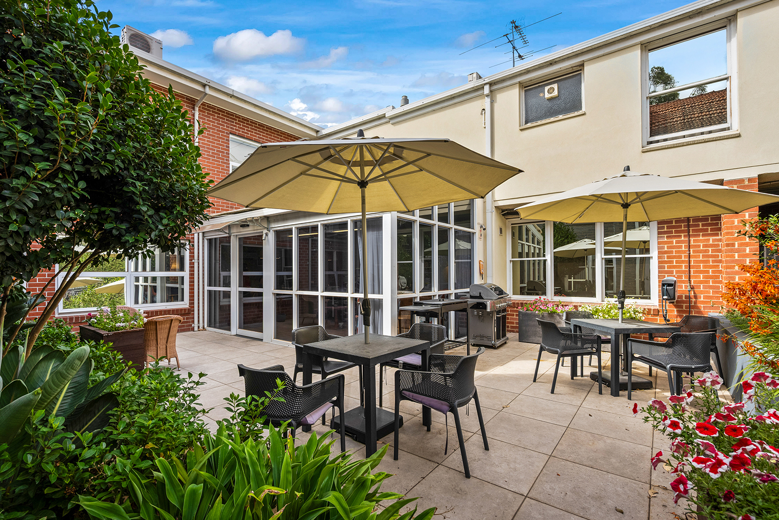 A courtyard with flowers in bloom and umbrellas attached to outdoor furniture.
