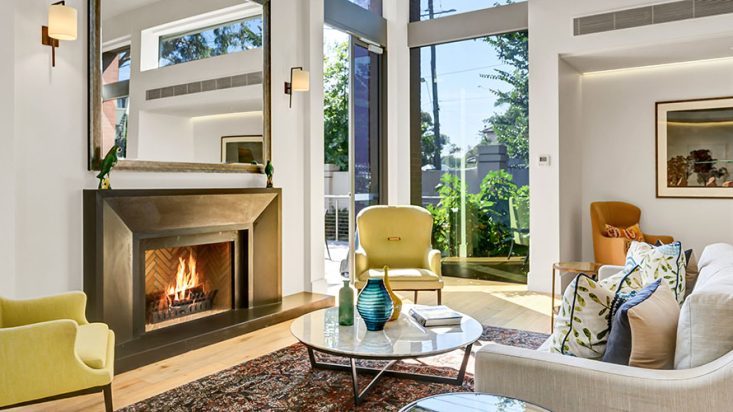 Yellow and grey couches sit around a coffee table, with gas fireplace on the wall. Trees can be seen through the window.