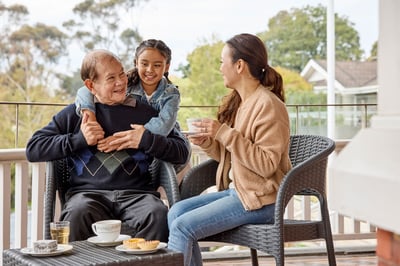 An older man sitting down in a chair. A young girl standing behind him with her arms wrapped around the man. Young woman sitting opposite the man, holding a teacup and saucer.
