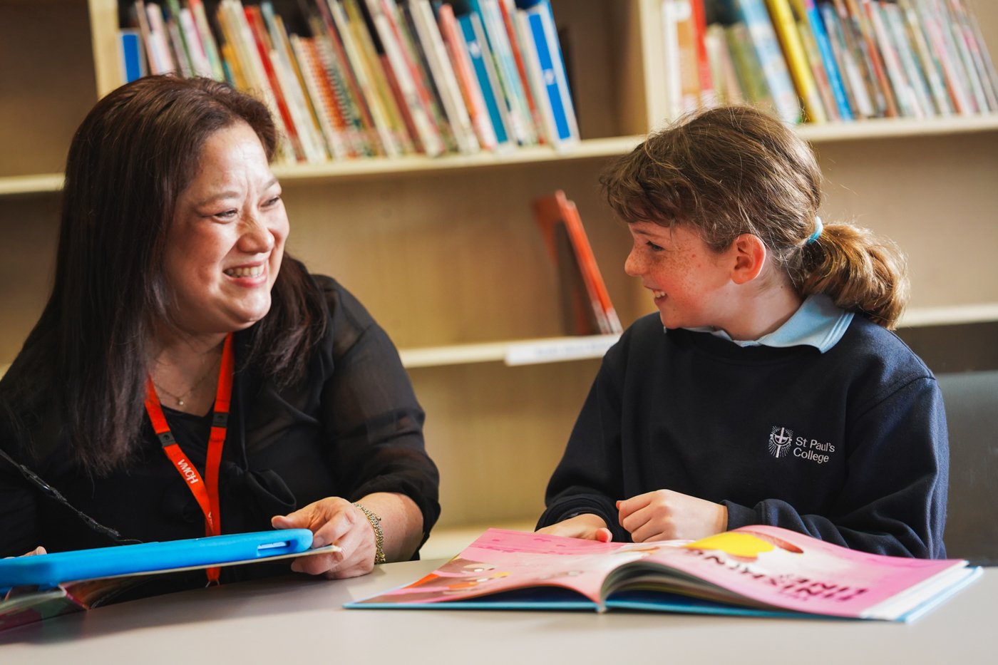 A teacher sitting at a table, holding a blue tablet device, smiling at a female student wearing a blue uniform with a pink book in front of her.